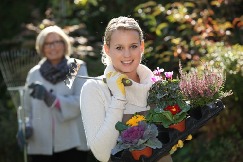 A young woman with blonde hair, wearing a white sweater and yellow gardening gloves, is smiling and holding a black tray filled with colorful flowering plants, including purple, yellow, and red blooms, in a well-maintained garden. In the background, an older woman with glasses and a scarf is seen, also engaged in gardening activities, holding a garden fork. The garden features lush green foliage, a variety of shrubs, and a neatly trimmed hedge, with dappled sunlight filtering through the trees, creating a bright and inviting outdoor environment suitable for gardening tasks typical of services provided by Lawn Mowing Forest Hill in the local area near Forest Hill, London.