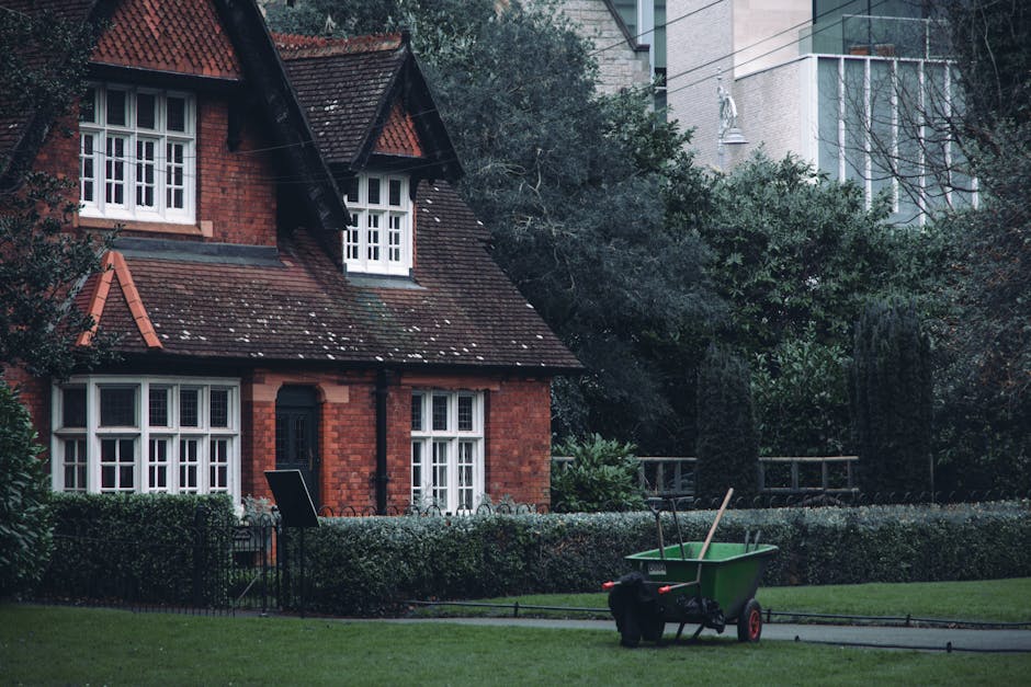 A quaint red-brick house with a steep, tiled roof and white-framed windows situated in a garden setting. The garden features well-maintained, trimmed hedges bordering a lush green lawn with a narrow p